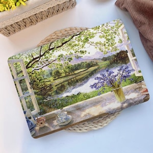 Puede incluir: Bandeja decorativa con una pintura de paisaje que representa un río y árboles vistos a través de una ventana. Un jarrón con flores moradas y una taza de té están en el alféizar. La bandeja tiene un asa tejida.