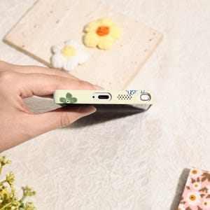 May include: A light green phone case with a floral design. The case features green leaves and blue accents. The phone case is held in a hand, with a blurred background of decorative flowers.