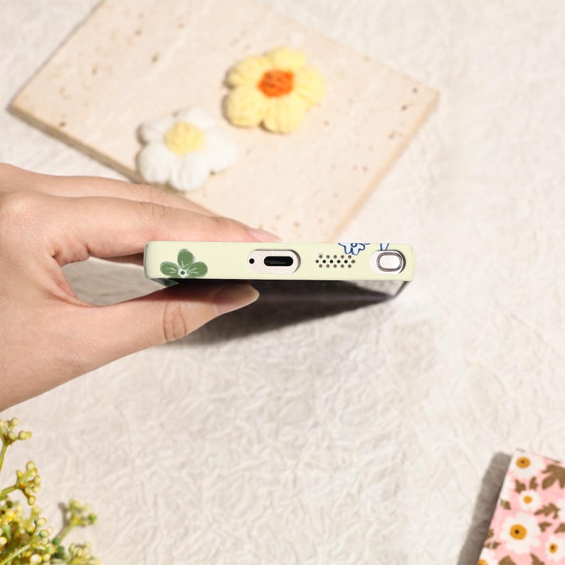 May include: A light green phone case with a floral design. The case features green leaves and blue accents. The phone case is held in a hand, with a blurred background of decorative flowers.