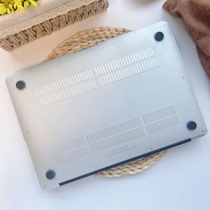 May include: A clear, frosted laptop case with ventilation slots and four black rubber feet. The case is designed to protect the bottom of a laptop. The case is sitting on a woven, circular mat.