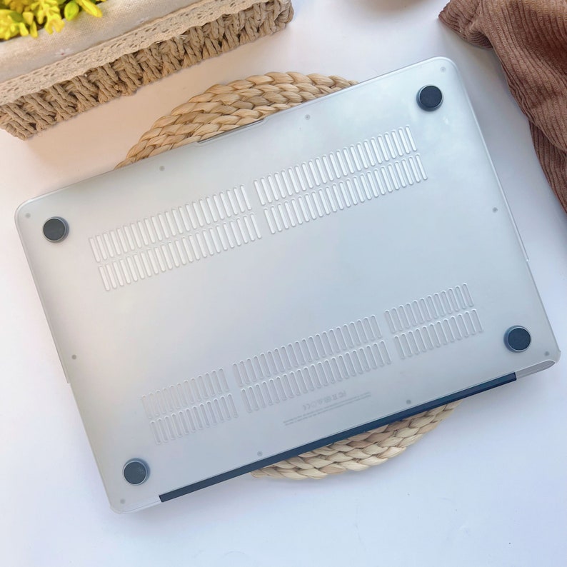 May include: A clear, frosted laptop case with ventilation slots and four black rubber feet. The case is designed to protect the bottom of a laptop. The case is sitting on a woven, circular mat.