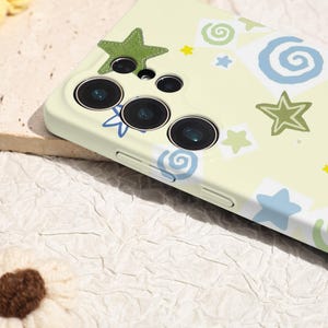 May include: A light yellow phone case with a whimsical design featuring green, blue, and yellow stars and swirls. The case has a camera cutout with three lenses and a flash. The case is made of a smooth material.