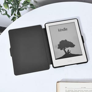 May include: A black e-reader case with a white Kindle e-reader inside. The e-reader screen shows a silhouette of a person sitting under a tree with the word "kindle" displayed on the screen.