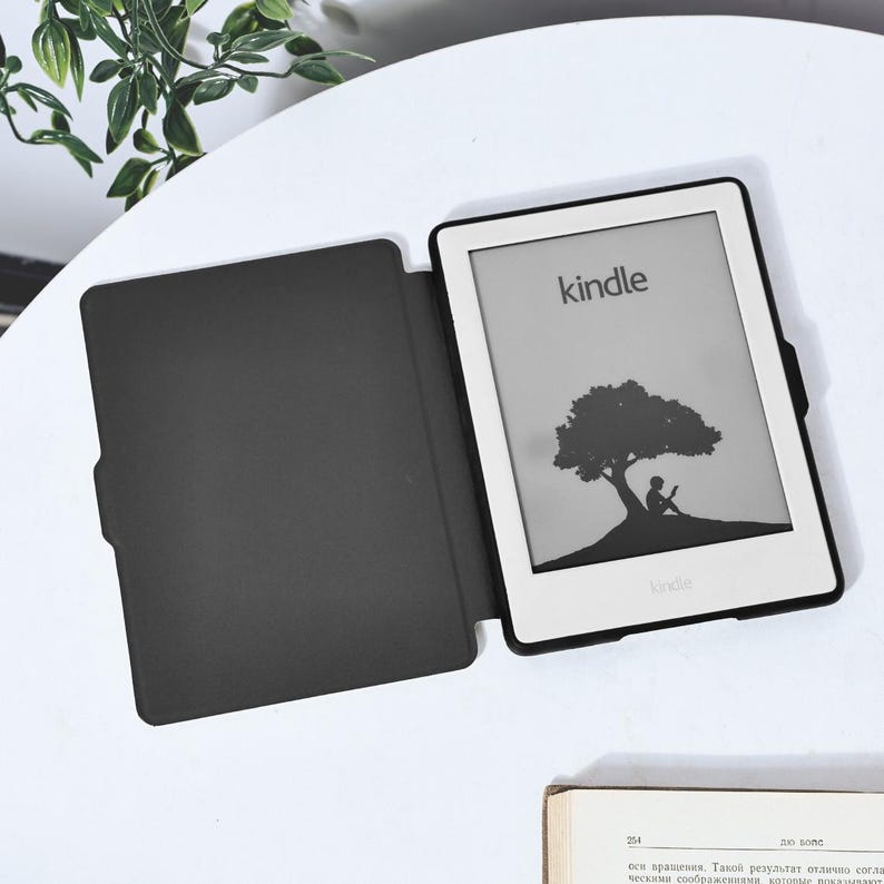 May include: A black e-reader case with a white Kindle e-reader inside. The e-reader screen shows a silhouette of a person sitting under a tree with the word "kindle" displayed on the screen.