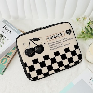 Könnte beinhalten: Eine schwarze und weiße karierte Laptoptasche mit braunem Hintergrund. Die Tasche zeigt eine Grafik mit zwei schwarzen Kirschen und dem Text "CHERRY." Der Text unter den Kirschen lautet: "But every once in a while You find someone who's iridescent And when you do Nothing will ever compare".