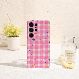 May include: A pink phone case with a gingham pattern and colorful star designs. The case has a camera cutout and is decorated with various star shapes in colors like blue, green, yellow, and silver.