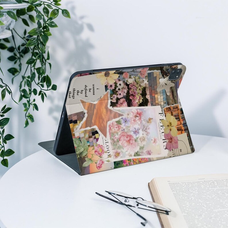 May include: A floral-themed tablet case with a collage design featuring a star, flowers, and text. The case is open, revealing the tablet screen. The design includes various colors, such as pink, yellow, and beige. The case is on a white table with a book and glasses.