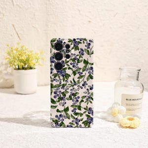 Op de afbeelding: Een telefoonhoesje met een patroon van aquarel blauwe bessen en groene bladeren op een witte achtergrond. De hoes heeft een camera aan de linkerbovenkant. De hoes ligt op een witte ondergrond.