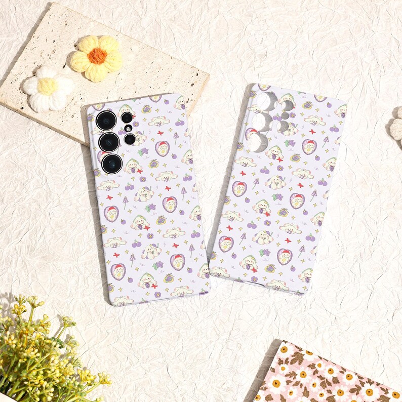 Może przedstawiać: Dwa etui na telefon z powtarzającym się wzorem postaci z kresk&oacute;wek, chmur, serc i innych małych ikon na jasnofioletowym tle. Etui mają błyszczące wykończenie i są przeznaczone do smartfona. Etui leżą na białej powierzchni.
