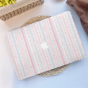 Puede incluir: Una funda para portátil con un diseño floral. La funda presenta rayas verticales en rosa claro, azul claro y blanco, adornadas con pequeñas flores rosas. El logotipo de Apple es visible.