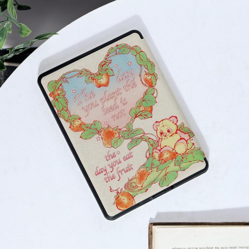 May include: A cream-coloured e-reader case with a black border. The case features a colourful illustration of a heart-shaped vine with strawberries, a bear, and the text "The day you plant the seed is not the day you eat the fruit."