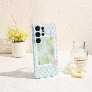 May include: A light blue phone case with a geometric pattern and a watercolor design. The case features a camera cutout and a decorative bow. The background includes a small potted plant and a glass bottle.