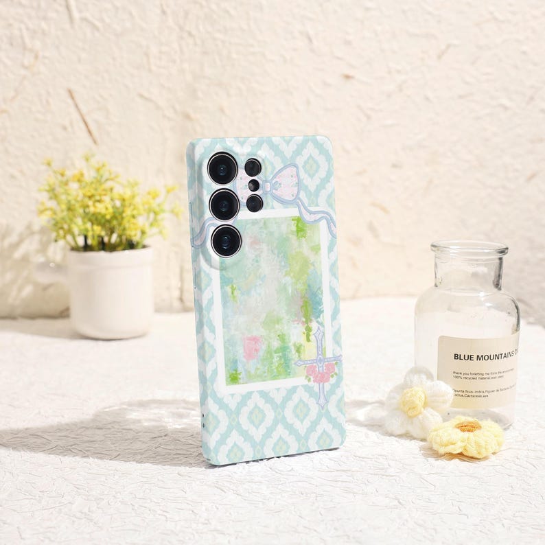 May include: A light blue phone case with a geometric pattern and a watercolor design. The case features a camera cutout and a decorative bow. The background includes a small potted plant and a glass bottle.