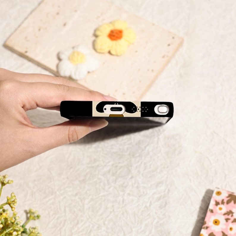 May include: Black phone case with a cartoon design. The case features a black border with a cream-colored design and a small, circular camera cutout. The design includes a cartoon face with a large, open mouth.