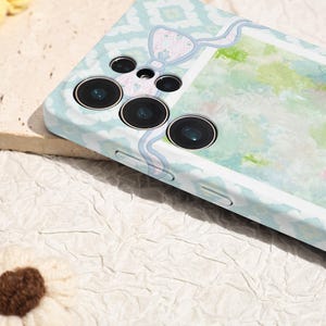 May include: A light blue phone case with a decorative bow and a watercolor design. The case has a patterned design and three camera lenses. The case is designed to protect the phone.