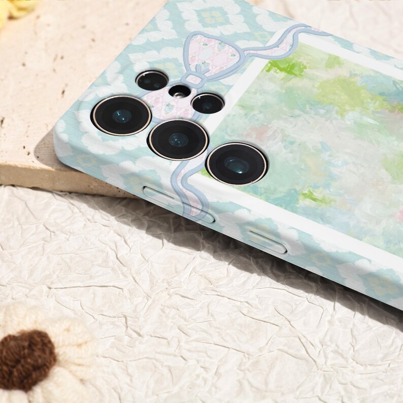 May include: A light blue phone case with a decorative bow and a watercolor design. The case has a patterned design and three camera lenses. The case is designed to protect the phone.