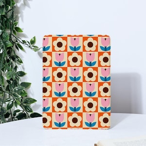 以下が含まれることがあります： 花と幾何学模様の繰り返し模様が施された長方形の装飾品。デザインは、ピンク色のチューリップのような花、茶色のデイジーのような花、青色のアクセントが、オレンジとベージュの正方形のグリッドに対して配置されています。白い表面に置かれています。