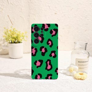 Puede incluir: Una funda de teléfono verde con un diseño de estampado de leopardo rosa y negro. La funda tiene un módulo de cámara con tres lentes. El fondo incluye una pequeña planta en maceta y una botella de vidrio.