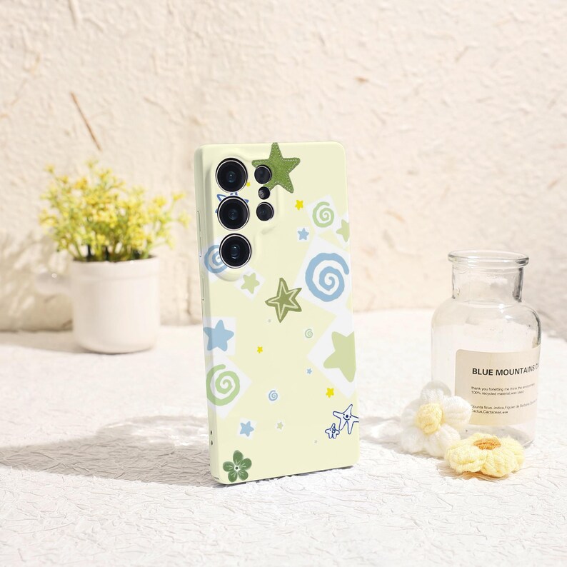 May include: A light yellow phone case decorated with green, blue, and yellow stars, swirls, and a flower. The case has a camera module with three lenses. The case is designed for a smartphone.
