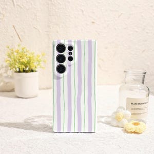 Puede incluir: Una funda de teléfono blanca con un patrón de rayas verticales en morado claro y verde. La funda tiene un recorte para la cámara con múltiples lentes. El fondo presenta una pequeña planta en maceta, una botella de vidrio y flores decorativas.