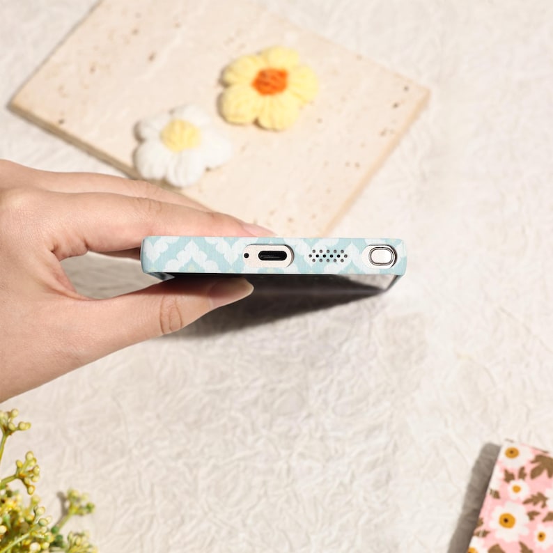 May include: A light blue phone case with a white geometric pattern. The case has a port for charging and a speaker. The case is held by a hand, with two decorative flower accents in the background.