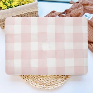 May include: A pink and white checkered laptop case with the Apple logo. The case is on a woven mat, with a basket of yellow flowers and a brown tote bag in the background.