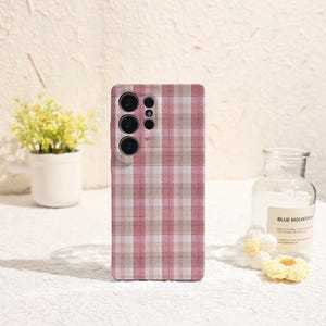 May include: A pink and white checked phone case with multiple camera lenses. The case has a rectangular shape and a grid pattern. The phone case is designed to protect the phone from scratches and damage.