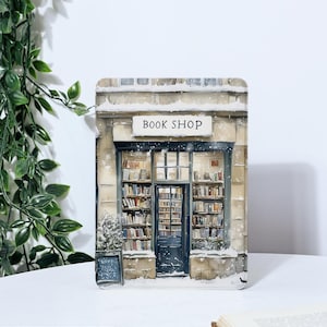 以下が含まれることがあります： 「BOOK SHOP」と書かれた看板のある本屋の店の水彩画。店は雪に覆われており、大きな窓には本がぎっしり並んでいます。