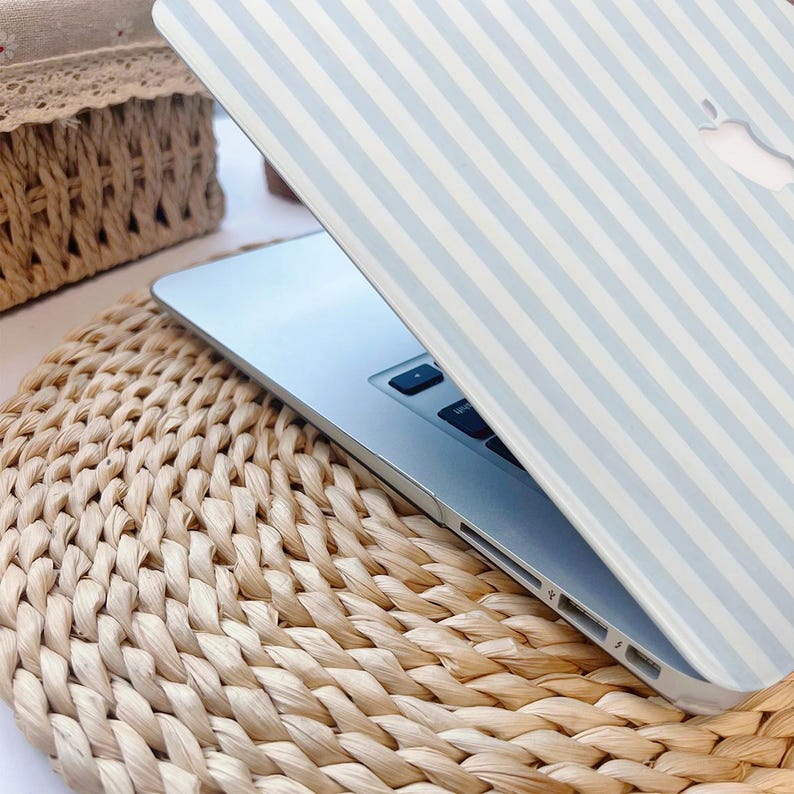 May include: A silver laptop with a light blue and white striped cover is open on a woven straw mat. The laptop's screen is partially visible, and the Apple logo is cut out of the cover. The mat is round and has a natural, textured appearance.
