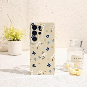 May include: A beige phone case adorned with a delicate floral pattern featuring white, blue, and navy blossoms, along with small brown leaves. The case has cutouts for the phone's camera and buttons, offering both style and protection.