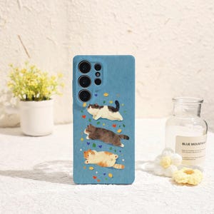 May include: A light blue smartphone case featuring a printed design of three cats in different colours—black and white, brown, and orange—positioned one above the other, surrounded by small colourful hearts, stars, and leaf-shaped icons.
