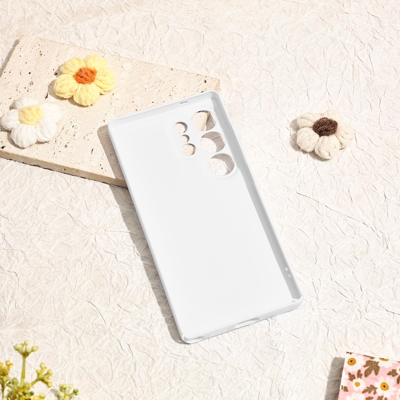 May include: A white phone case with cutouts for the camera and buttons. The case is surrounded by handmade crochet flowers in yellow, white, and brown, set against a textured, off-white background.