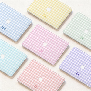 May include: A collection of laptop cases in various pastel colors, including blue, yellow, pink, green, and purple. Each case features a gingham pattern and a personalized monogram. The Apple logo is visible on each case.