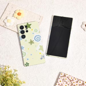 May include: Two phone cases. One is light green with a pattern of stars, swirls, and a small airplane. The other is a black screen with a light green border. Crochet flowers are in the background.