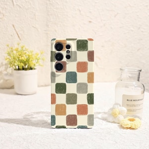 Può includere: Una custodia per telefono con sfondo crema e un motivo ripetuto di quadrati colorati nelle tonalità di verde, arancione, marrone e grigio. La custodia ha un modulo fotocamera con tre obiettivi.