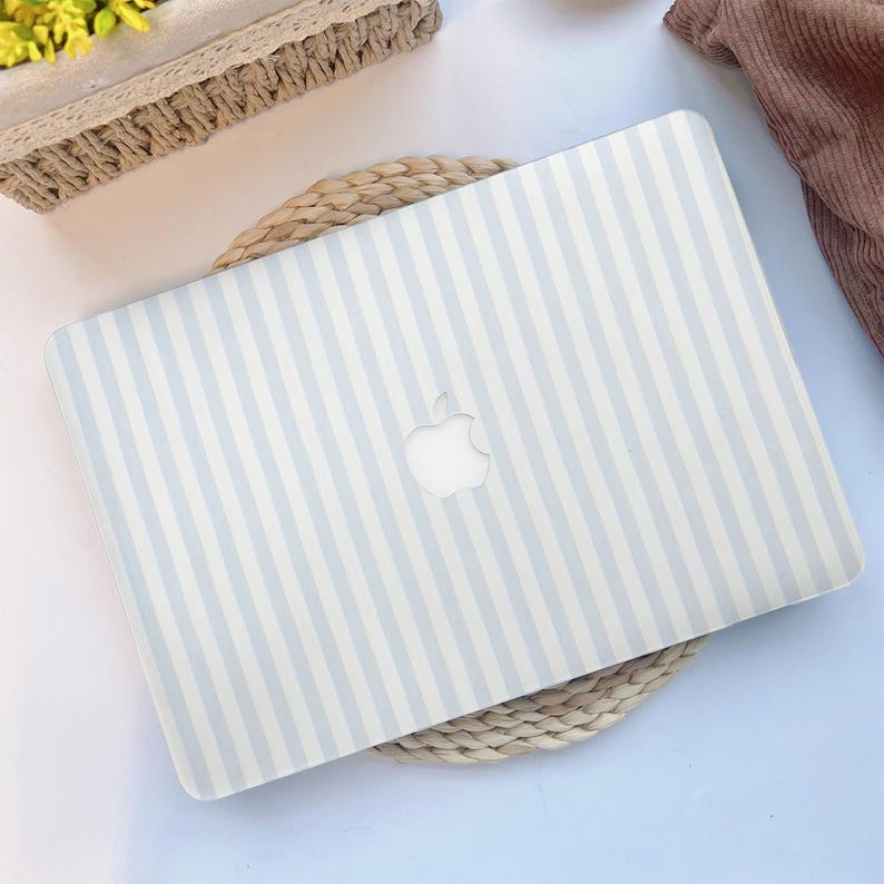 May include: A light blue and white striped laptop case with the Apple logo cutout. The case is rectangular and covers the top of the laptop. The case is resting on a woven circular mat.