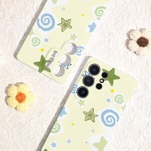 May include: Two light yellow phone cases with a whimsical design. The cases feature green stars, blue swirls, and yellow star accents. One case has a green star applique. The cases are on a textured white surface.
