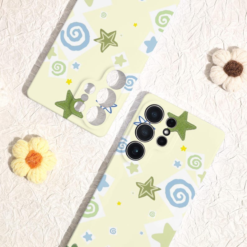 May include: Two light yellow phone cases with a whimsical design. The cases feature green stars, blue swirls, and yellow star accents. One case has a green star applique. The cases are on a textured white surface.