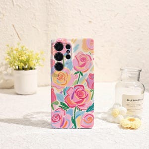 Puede incluir: Una funda de teléfono con un diseño floral con rosas rosas, amarillas y azules sobre un fondo blanco. La funda tiene un módulo de cámara con tres lentes y un flash. El estilo general es artístico y colorido.