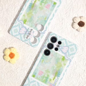 May include: Two light blue phone cases with a pastel watercolor design. Each case features a bow and flower embellishment. The cases have a patterned background and a rectangular frame. One case has a camera array.