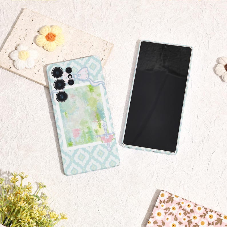 May include: A light blue phone case with a white and blue geometric pattern and a watercolor design. The case has a bow and a floral design. A second phone is next to the case.