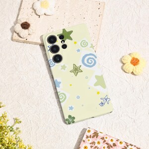 May include: A light yellow phone case with a whimsical design featuring green stars, blue and green swirls, and small yellow stars. The case has a multi-lens camera cutout and is surrounded by decorative flowers.