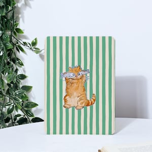 May include: A rectangular notebook with a green and white striped pattern. The cover features a watercolor illustration of a ginger cat holding two fish in its mouth. The cat is sitting upright.