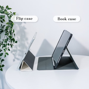 May include: Two tablet cases, a flip case and a book case, are displayed on a white table. The flip case is clear with a gray and white base. The book case is black with a gray base. The text "Flip case" and "Book case" are above each case.
