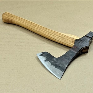 Könnte beinhalten: Eine handgeschmiedete Axt mit einem Holzstiel. Der Axtkopf ist aus Stahl und hat eine gebogene Klinge. Der Stiel ist aus hellem Holz und hat eine natürliche Oberfläche.