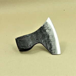 Könnte beinhalten: Ein schwarzer Axtkopf mit einer leicht gebogenen Klinge. Die Klinge ist poliert und hat eine hellgraue Oberfläche. Der Axtkopf ist aus Metall und hat eine strukturierte Oberfläche.