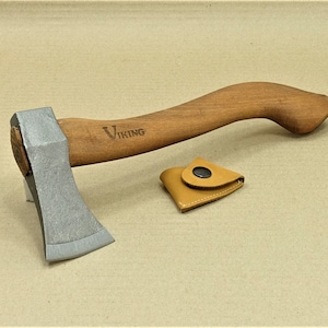 Könnte beinhalten: Eine kleine Axt mit einem Holzstiel und einer Lederscheide. Der Axtkopf ist silberfarben und der Stiel ist braun. Die Scheide ist aus braunem Leder mit einem schwarzen Knopf.