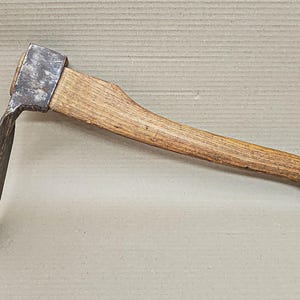 May include: A vintage hoe with a wooden handle and a metal blade. The handle is made of light brown wood, and the blade is a dark, weathered metal. The tool is designed for agricultural use, likely for weeding or cultivating soil.