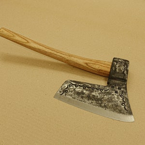 Peut inclure: Une hache forgée à la main avec un manche en bois et une lame incurvée décorative. La lame présente un motif de petits cercles et de lignes.