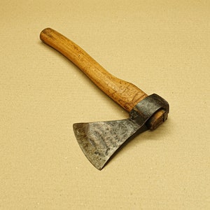 Könnte beinhalten: Eine kleine Axt mit einem Holzstiel und einem Metallkopf. Die Axt hat einen braunen Holzstiel und einen dunkelgrauen Metallkopf. Die Axt liegt auf einer braunen Oberfläche.